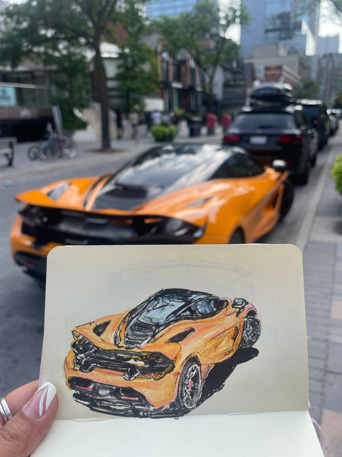 A hand displays a sketchbook showcasing a hand-drawn illustration of an orange sports car that perfectly mirrors the real vehicle parked on the street behind it. With urban buildings and trees framing the background, this scene highlights how a Custom Car Portrait Commission can capture any car in a personalized and artistic way—making it an ideal gift for car enthusiasts.