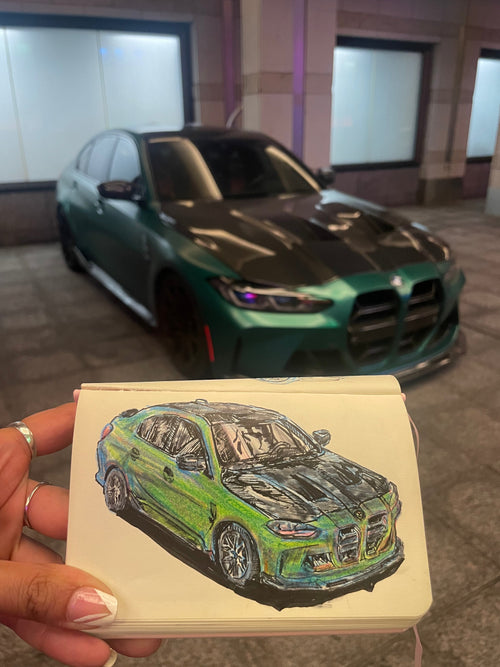 A hand holds a sketchbook featuring a Custom Car Portrait Commission of a green and black BMW, perfectly matching the real car parked behind it on a city street at night—an impressive piece of hand-drawn automotive art, making it an ideal gift for any car enthusiast.